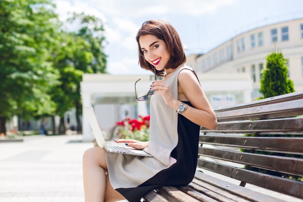 회색과 검은 색 드레스를 입은 예쁜 갈색 머리 소녀가 도시의 벤치에 앉아 있습니다. 그녀는 무릎에 노트북을 들고 카메라에 친절하게 웃고 있습니다.