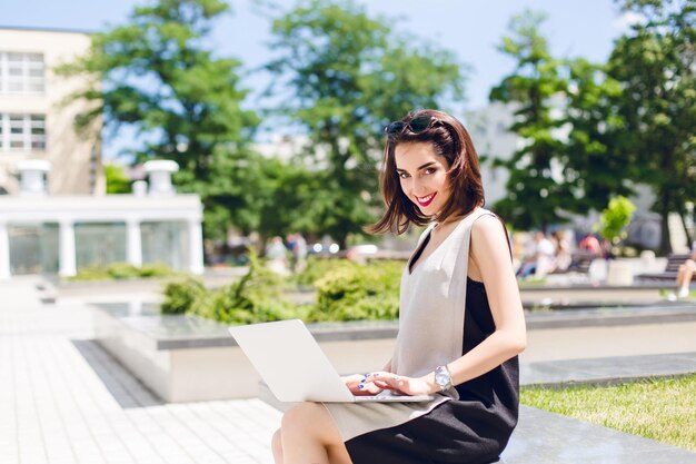 A pretty brunette girl in gray and black dress is sittting in the park in city. She is typing on laptop and smiling with vinous lips to the camera.