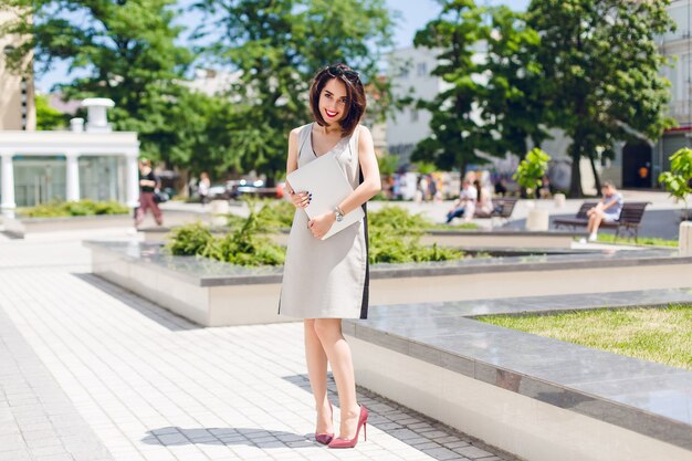 Pretty brunette girl in gray dress and vinous heels is standing in park in the city. She holds laptop and looks shy.