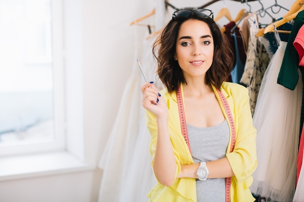 Una bella ragazza mora con un vestito grigio e una giacca gialla è in piedi vicino ai vestiti nello studio del laboratorio. tiene una matita in mano e sorride alla telecamera.