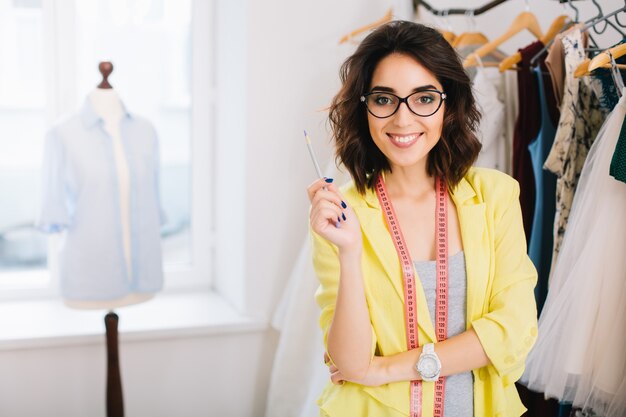 A pretty brunette girl in a grey dress and yellow jacket is standing near clothes in workshop studio. She is  smiling to the camera.