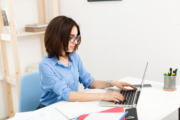 A pretty brunette girl is sitting and typing on laptop at the table in office. She wears blue shirt and black glasses.