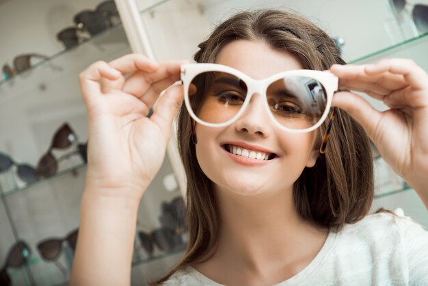 Красивая брюнетка девушка улыбается, примеряет солнцезащитные очки в магазине оптики