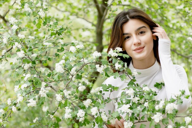 Pretty brunette girl standing in tree blossom looking at camera smiling touching hair