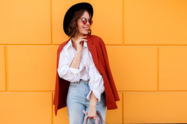 Ragazza graziosa del brunette in cappello alla moda che propone sopra la parete gialla.