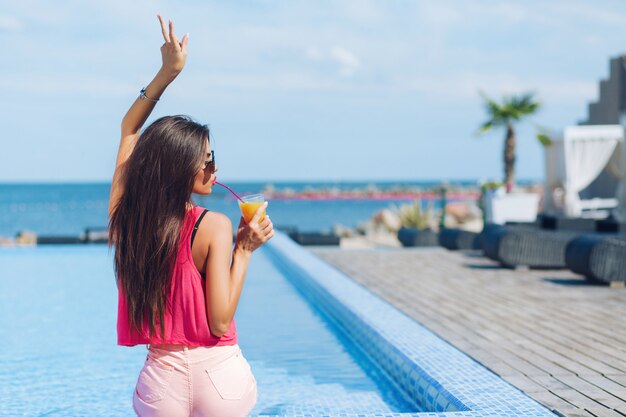 Pretty brunette girl with long hair is sitting near pool. She holds hand above and drinks through a straw. View from back.