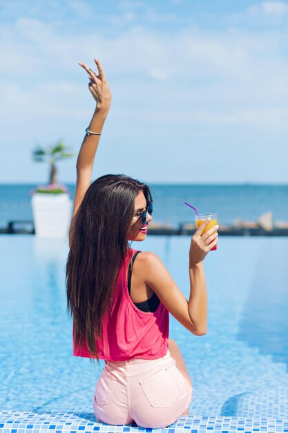 Pretty brunette girl with long hair is sitting near pool. She wears rose T-shirt, shorts, sunglasses. She holds drink in one hand and another one above. View from back.