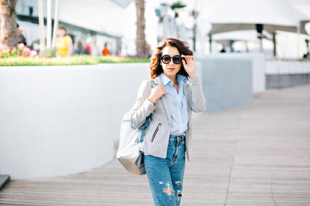 Симпатичная брюнетка с короткими волосами позирует перед камерой в городе. Она носит рубашку, джинсы, куртку и сумку. Она держит солнцезащитные очки на лице и смотрит в камеру.