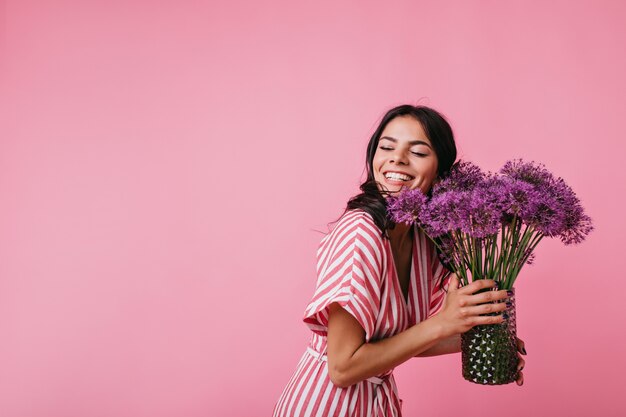 きれいなブルネットは、提示された花を楽しみながら、まばゆいばかりの笑顔です。幸せから目を閉じているピンクの縞模様のトップの女の子の肖像画。