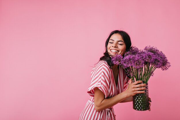 Pretty brunette is dazzlingly smiling while enjoying presented flowers. Portrait of girl in pink striped top closing her eyes from happiness.