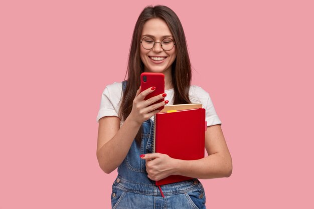 Pretty brunette woman holds mobile phone, sends text message, happy to read comments under post, holds textbook