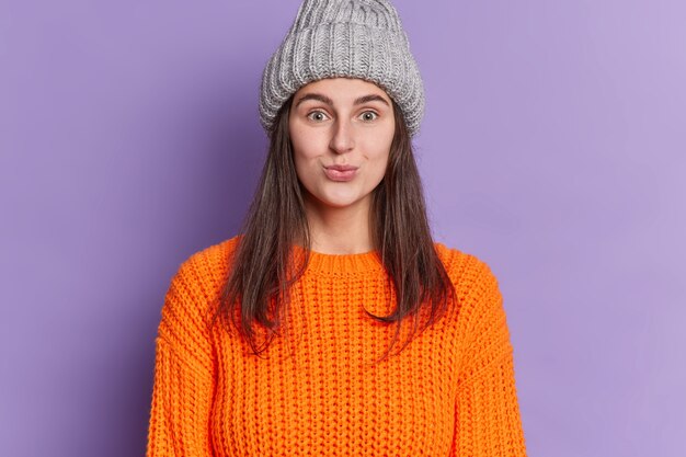 Симпатичная брюнетка женщина выглядит с забавным выражением лица, надувает губы, носит вязаную серую шляпу, оранжевый свитер, наслаждается зимним сезоном.