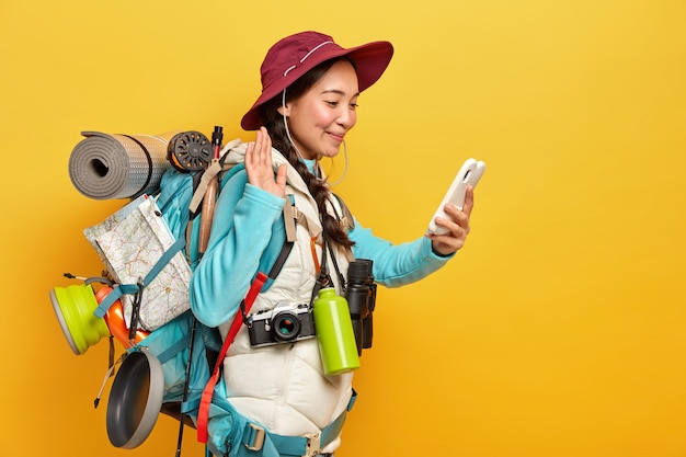 Pretty brunette woman makes video call, waves with palm at smartphone camera, uses modern technology for keeping in touch with friends during expedition