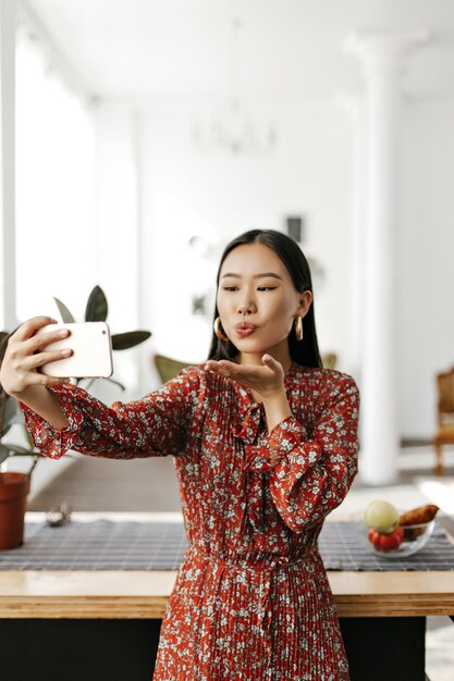 Pretty brunette woman in red dress blows kiss and takes selfie in cozy room Charming Asian lady in floral outfit holds phone