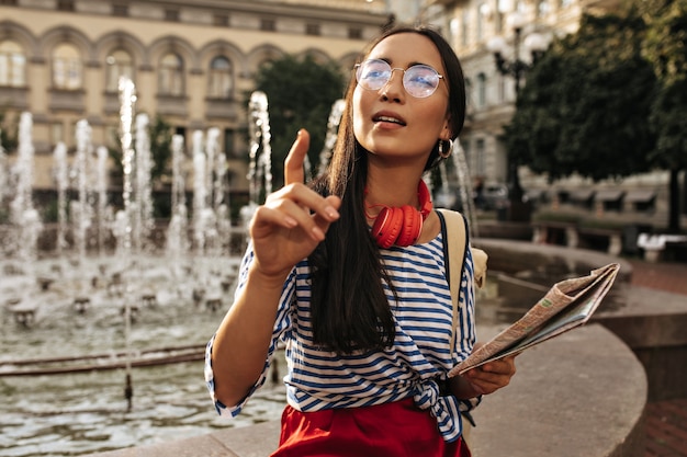 Pretty brunette woman in stylish striped shirt, red headphones and silk skirt points into distance