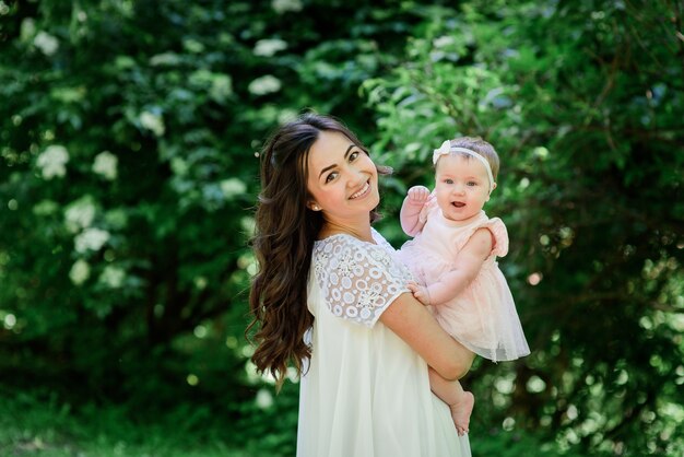 Pretty brunette woman in white dress poses with her little daughter in the garden