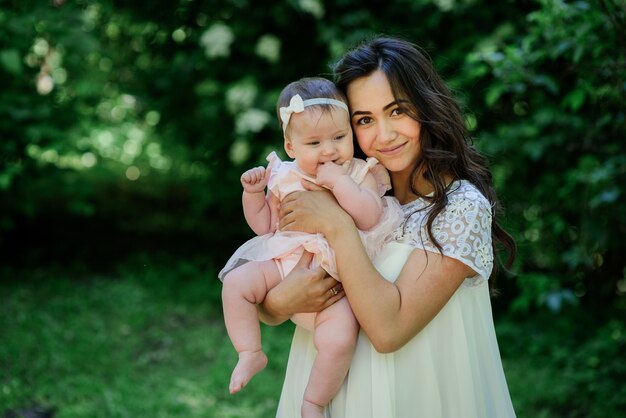 Pretty brunette woman in white dress poses with her little daughter in the garden