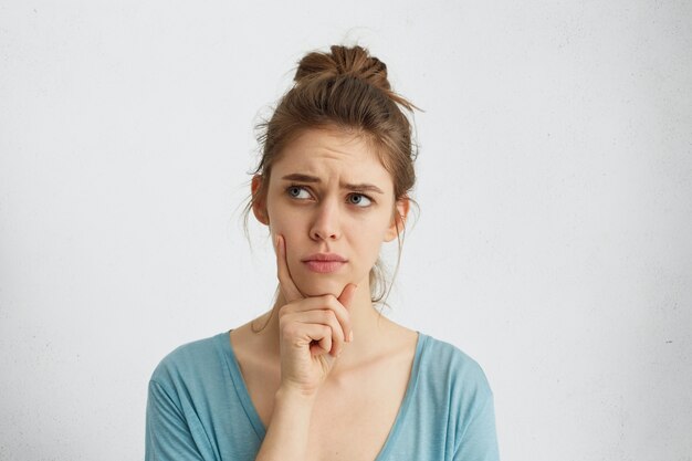 Pretty brunette woman with blue eyes and pinned-up hair frowning her eyebrow while looking aside with placid and thoughtful look. Pensive female with puzzled expression thinking something over