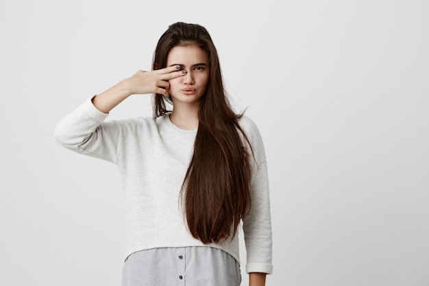 Pretty brunette woman with long hair, making faces, pouting lips, closing one of her eyes with v sign, having pleased expression. Beautiful young female dressed in casual clothes posing indoors