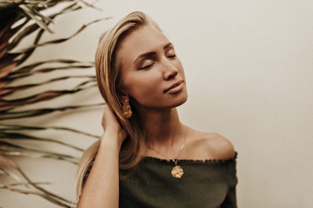 Free photo pretty calm young tanned blonde woman in dark green top with golden medallion poses with closed eyes near white wall with palm leaf