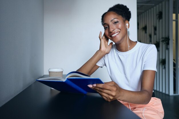 Pretty casual African American girl in earphones with book and coffee joyfully looking in camera in modern co-working space