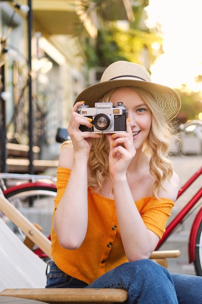모자를 쓴 꽤 캐주얼한 금발 소녀가 거리 카페의 갑판 의자에 있는 복고풍 카메라로 사진을 꿈꾸며 사진을 찍고 있습니다.
