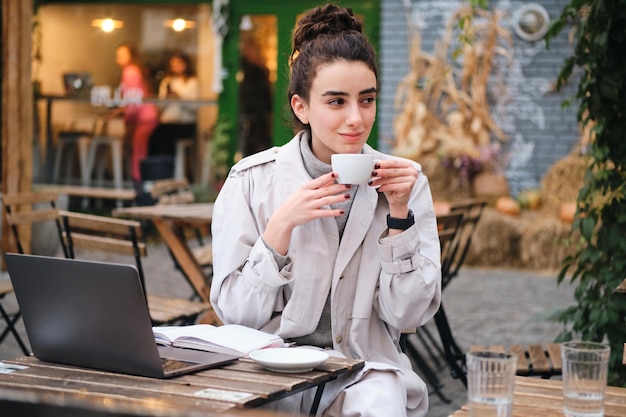 Pretty casual brunette girl in trench coat drinking coffee studying on laptop in cafe on city street