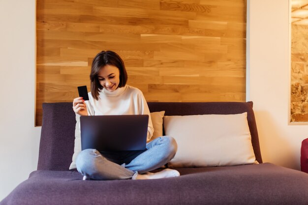 Pretty casual dressed woman in blue jeans and white t-shirt is working on laptop computer sitting on dark bed in front of wooden wall at home