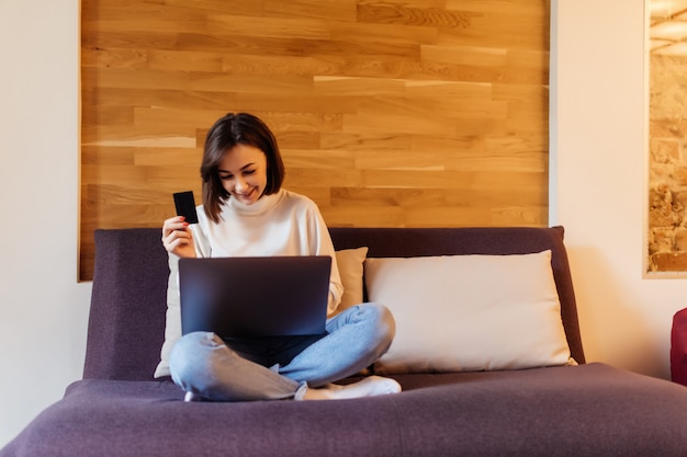Довольно повседневная одетая женщина в синих джинсах и белой футболке работает на ноутбуке, сидя на темной кровати перед деревянной стеной дома