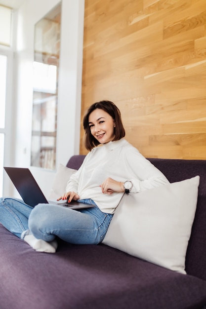Pretty casual dressed woman in blue jeans and white t-shirt is working on laptop computer sitting on dark bed in front of wooden wall at home