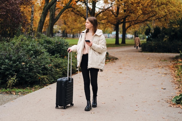 Pretty casual girl in down jacket thoughtfully walking through city park with suitcase