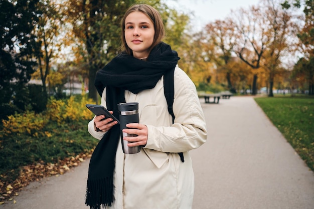 Pretty casual girl in down jacket walking around park alone with cellphone and thermo cup