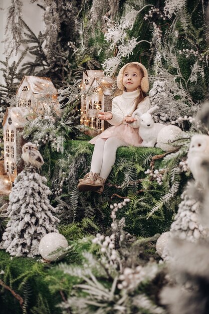 긴 머리를 가진 예쁜 백인 아이는 미소를 짓고 그녀 주위에 많은 장식 나무와 작은 토끼가 있는 크리스마스 분위기에 앉습니다.