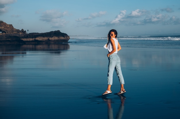 Pretty caucasian fit woman in white top and jeans on reflecting beach by ocean at sunset light