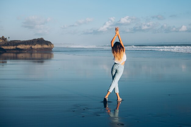 Pretty caucasian fit woman in white top and jeans on reflecting beach by ocean at sunset light