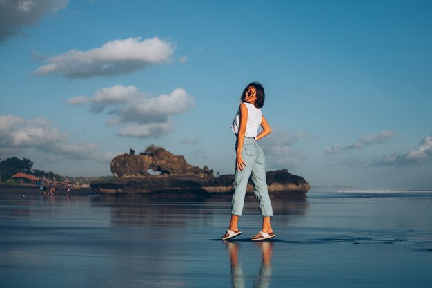Довольно кавказская женщина в белом топе и джинсах на отражающемся пляже у океана в лучах заката