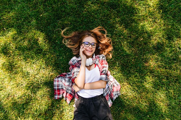 Pretty caucasian girl in glasses lying on green lawn. Overhead outdoor portrait of pleasant young woman chilling in park.
