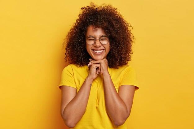 Симпатичная афро-девушка с кудрявыми волосами радуется жизни, держит руки под подбородком, чувствует себя вне себя от радости и удовлетворения, имеет естественную внешность, носит ярко-желтую футболку, позирует в помещении. Концепция счастья