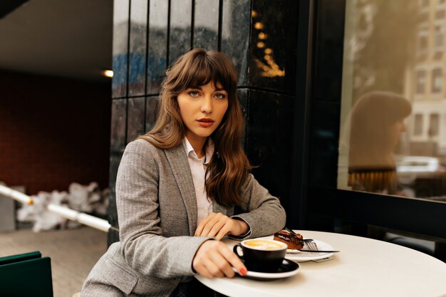 Pretty charming lady in grey jacket with long wavy hair sitting on city cafeteria is having coffee break