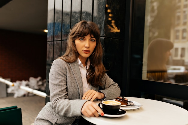 Pretty charming lady in grey jacket with long wavy hair sitting on city cafeteria is having coffee break