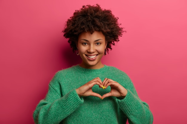 Una donna piuttosto affascinante modella il gesto del cuore, mostra cosa significa il fidanzato per lei, esprime affetto e amore, sorride piacevolmente, indossa un maglione verde, isolato su un muro rosa. concetto di linguaggio del corpo