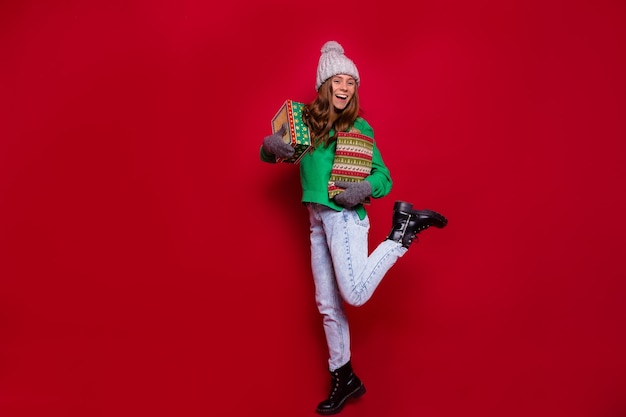 Pretty charming young woman with christmas gift box posing on red wall in winter outfit