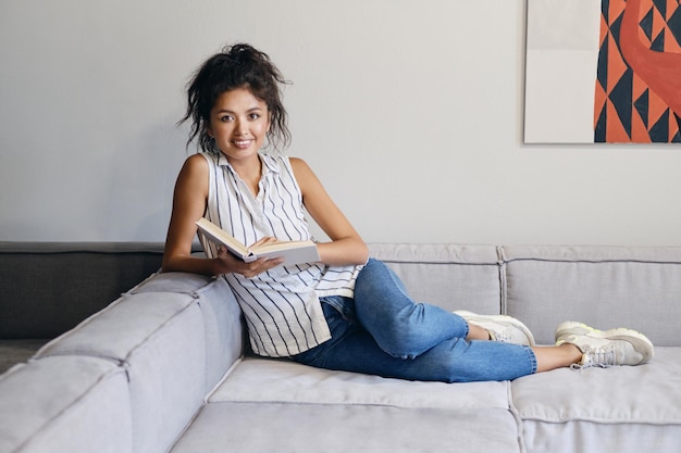 Pretty cheerful Asian girl lying on sofa with book joyfully looking in camera at modern home