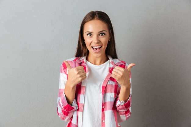 Pretty cheerful brunette woman in checkered shirt, showing thumbs up gesture