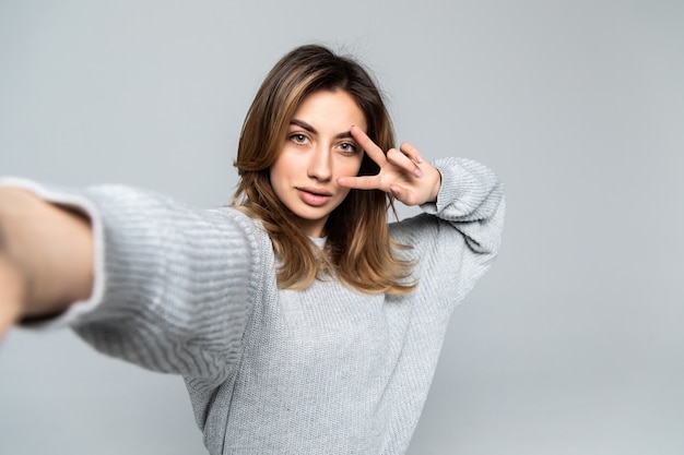 Pretty cheerful brunette woman showing peace gesture while taking selfie on smartphone, isolated over gray wall