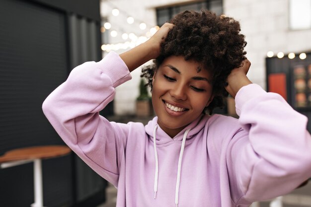 Pretty cheerful curly brunette darkskinned African woman in oversized purple hoodie smiles and touches hair outside