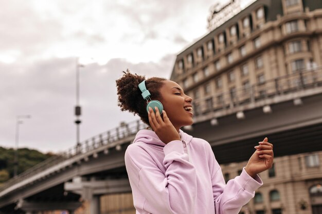 Pretty cheerful curly darkskinned lady in pink hoodie sings Attractive woman listens to music in headphones outdoors