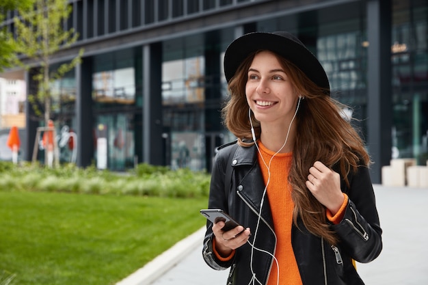 Pretty cheerful European girl in trendy wear, walks across metropolis, enjoys online playlist song in earphones