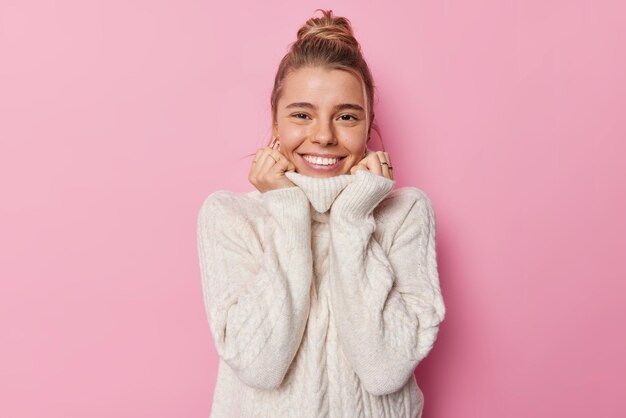 Pretty cheerful fair haired woman dressed in comfortable white knitted sweater feels warm and cozy smiles gladfully isolated over pink background People positive emotions and feelings concept