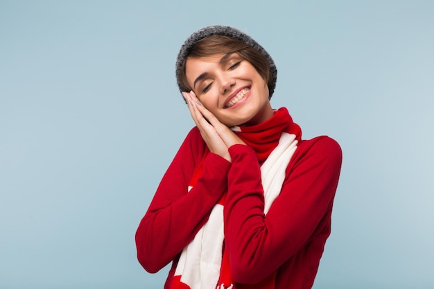 Pretty cheerful girl in red sweater, scarf and knitted hat dreamily closing eyes while showing sleepy gesture with hands over blue background